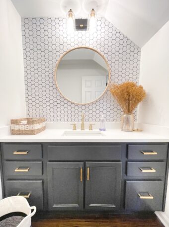 Black Panther by Benjamin Moore bathroom vanity, white quartz countertops, brass pulls and faucet, white hexagon backsplash with dark gray grout, wooden round mirror, 2 light sconce with clear glass cylindrical shades, woven basket decor, yellow babys breath in gold opaque vase